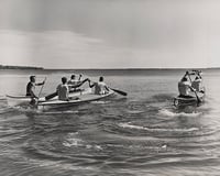 Rowers in three canoes paddle with oars in their hands across a lake in the middle ground, with the horizon and sky in the background.